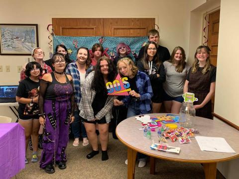 A group of teenagers posing together at a party. They are smiling and standing in front of a colorful backdrop. One person in the front holds a sign that says "90s."